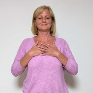Frau mit Hände am Brustbein - SeelenTON Akademie Woman with eyes closed, hands over chest, wearing a pink sweater, practicing deep breathing indoors.
