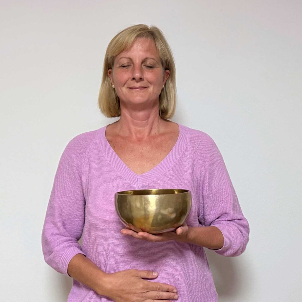 Woman with blonde hair in a pink sweater, eyes closed, holding a brass singing bowl at chest level in a meditative pose.