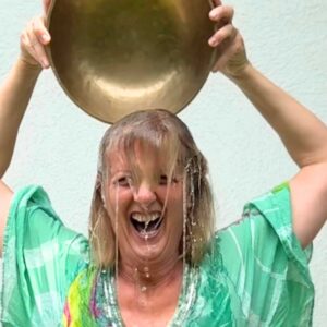 Person tilts a large bowl over their head while water pours down, smiling in a green striped shirt.