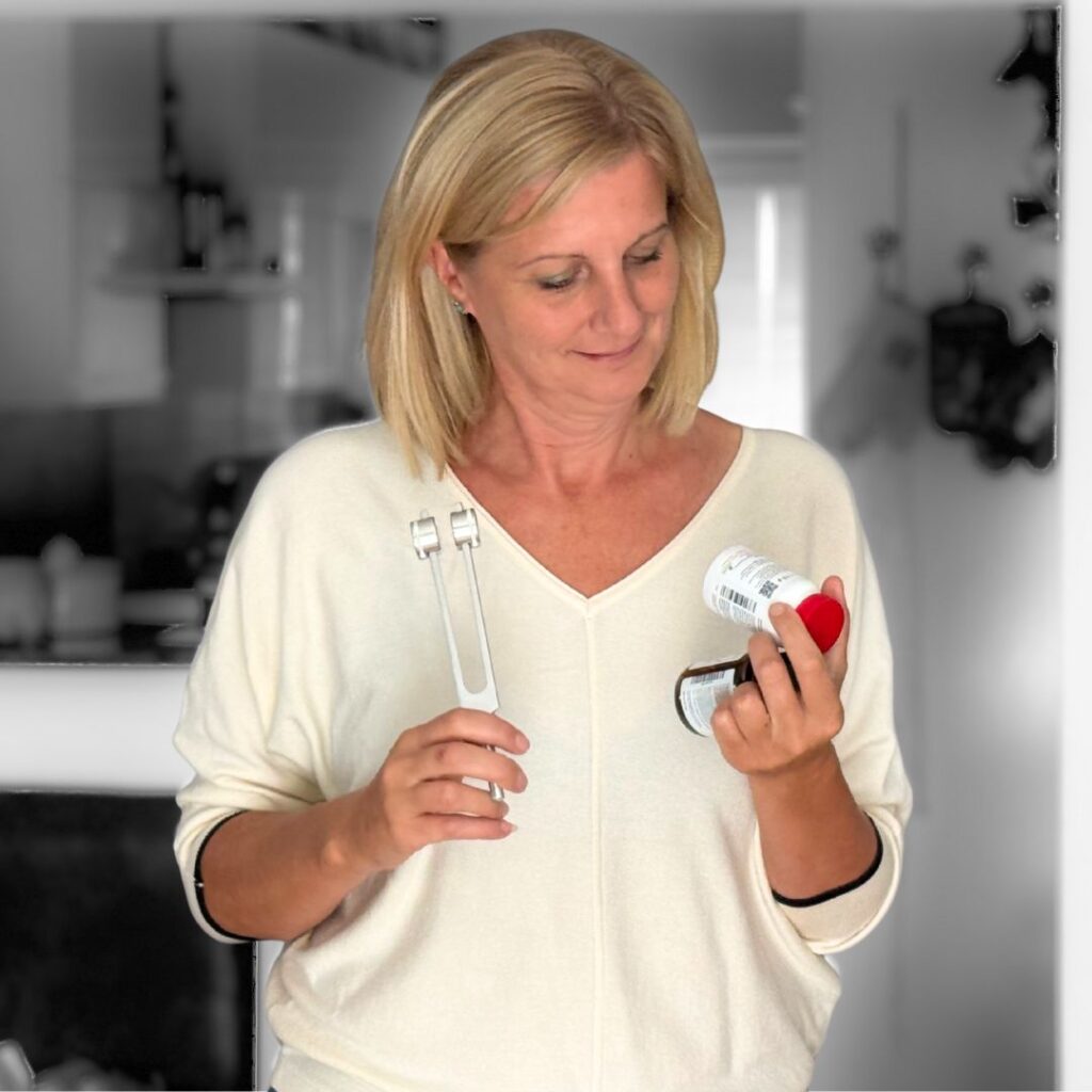 Woman with blond hair in a white sweater holds two prescription bottles and a kitchen tongs in a bright kitchen setting.